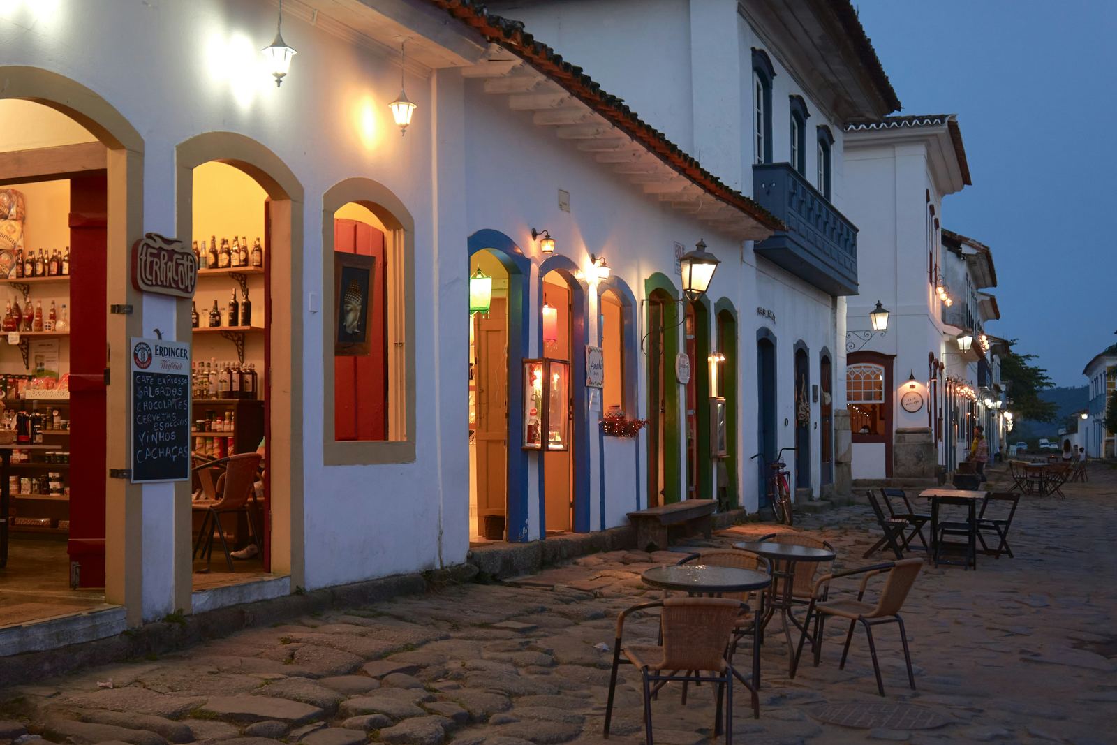 Rua do centro histórico ao anoitecer, com mesas na calçada e lampiões acesos