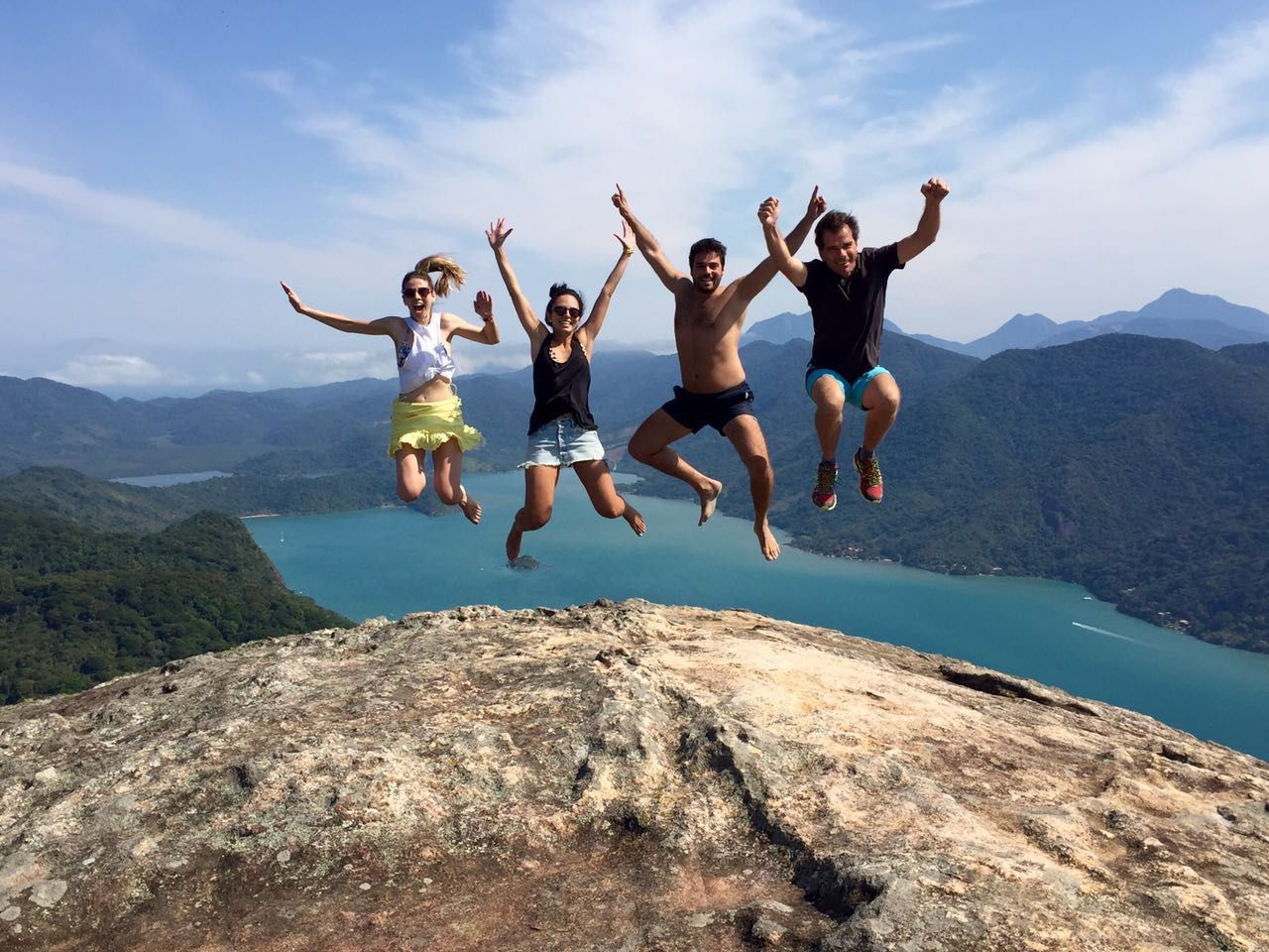 Quatro amigos pulando no topo do Pão de Açúcar do Mamanguá, com o braço de mar e as montanhas ao fundo