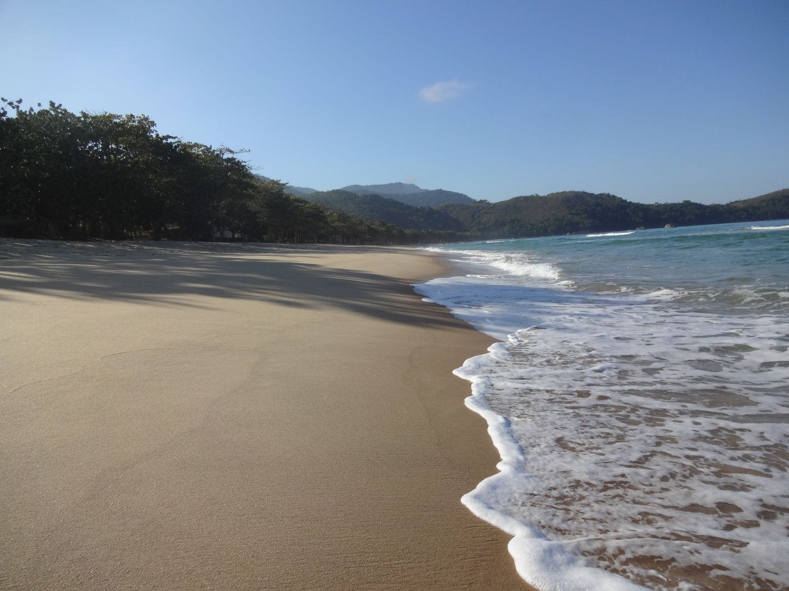 Longa praia de areia dourada com ondas e morros cobertos de mata